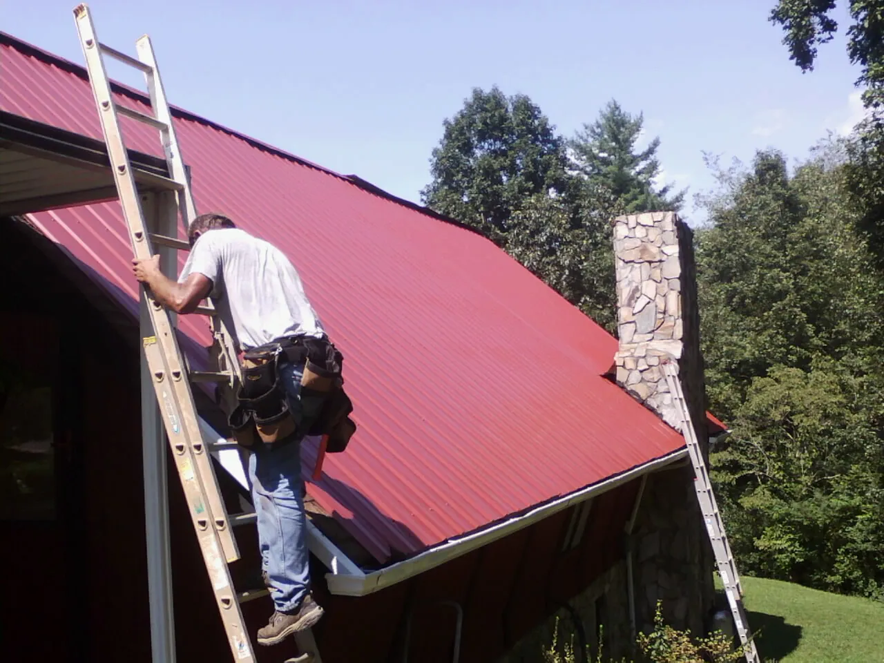 Roofer on a ladder beside a red metal roof and stone chimney — Soffit & Fascia Repair by Roofing Brothers in Elkton
